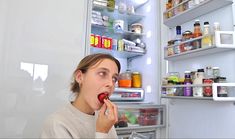 a woman is standing in front of an open refrigerator and eating something out of her hand