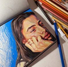 a pencil drawing of a man's face with his hand on his chin, next to colored pencils and crayons