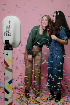 two women standing next to each other with confetti in front of them on a pink background