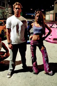 two people standing next to each other in front of a pink car and some buildings