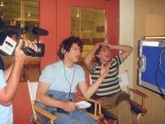 two men sitting in chairs with headphones on, and one man holding a camera