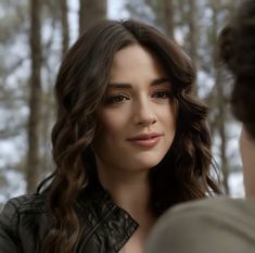 a woman with long brown hair is looking at another person in the woods and has her hand on her shoulder