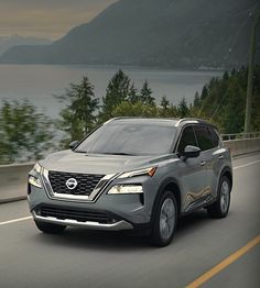 the front end of a silver nissan suv driving down a road next to trees and water