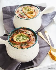 two white bowls filled with soup on top of a blue napkin next to gold spoons