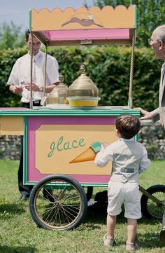 a little boy standing next to an old fashioned ice cream cart with a man behind it