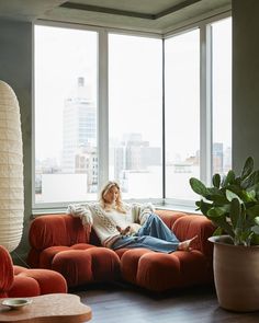 a woman sitting on top of a red couch in front of a window next to a potted plant