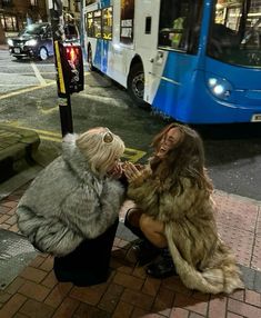 two women are sitting on the sidewalk near a bus and one is talking to another woman