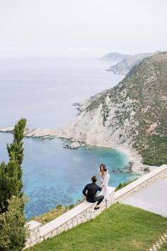 a man kneeling down next to a woman on top of a lush green hillside near the ocean