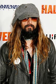 a man with long hair wearing a hat and scarf is standing in front of a wall