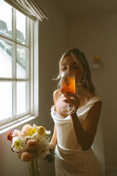 a woman in a white dress holding a wine glass next to a vase filled with flowers