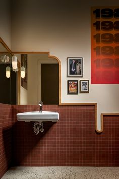 a sink and mirror in a bathroom with red tiles on the wall, framed pictures above it