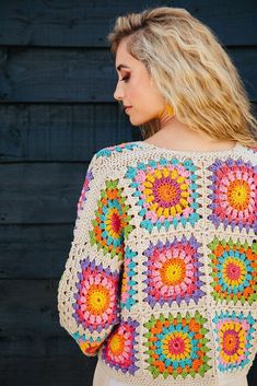 a woman wearing a multicolored crochet granny granny sweater standing in front of a wooden wall