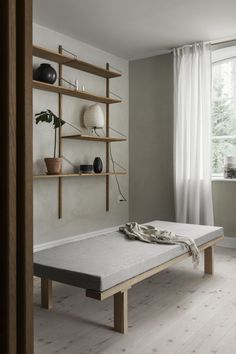 a bed sitting in front of a window next to a wooden shelf filled with vases