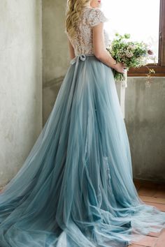 a woman standing in front of a window wearing a blue dress and holding a bouquet