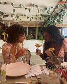 two women sitting at a table with wine glasses in their hands and flowers on the table