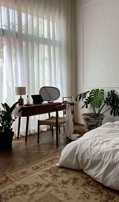 a bedroom with a bed, desk and plants in the corner on either side of the window