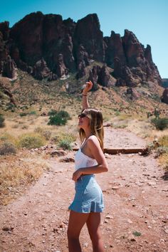 a woman standing in the middle of a dirt road with mountains and rocks behind her