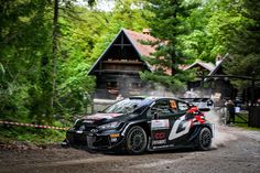 a black car driving down a dirt road next to a wooden building and forest covered hillside