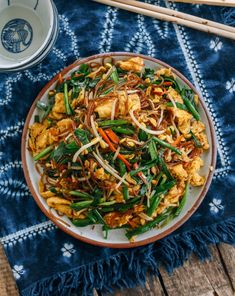 a white plate topped with stir fry vegetables next to chopsticks on a blue and white table cloth