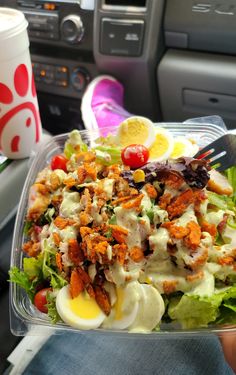 a salad in a plastic container with dressing and eggs on top, sitting next to a cup