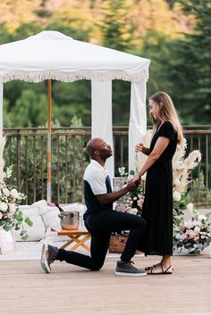 a man kneeling down next to a woman in front of a white tent with flowers