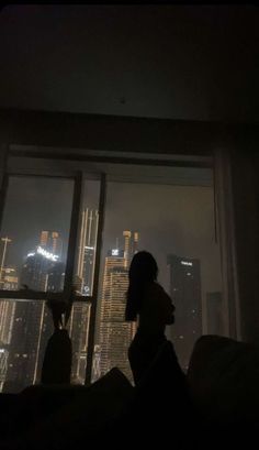 a woman standing in front of a window with the city lights lit up behind her