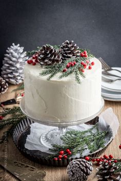 a white frosted cake with pine cones and red berries on top, sitting on a wooden table