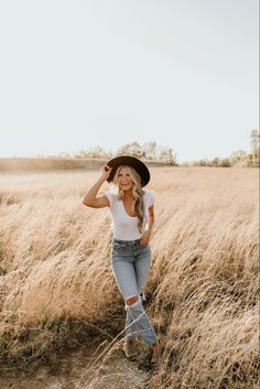 a woman standing in a field wearing a black hat and jeans with her hands behind her head