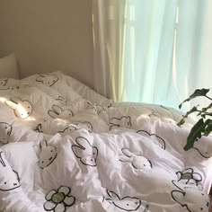 an unmade bed with white sheets and black drawings on it, next to a plant