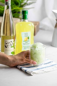 a hand holding a small cup filled with green liquid next to two bottles of alcohol