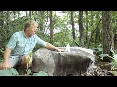 a man sitting next to a large rock in the middle of a forest with water coming out of it