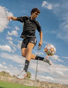 a young man kicking a soccer ball on top of a green field with clouds in the background