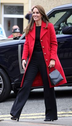 a woman in a red coat and black pants is walking down the street with her handbag