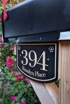 a black and gold house number sign attached to a wooden post with pink flowers in the background