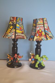 two lamps are sitting on a table with comic themed fabric coverings, one is turned on and the other has an iron man figurine