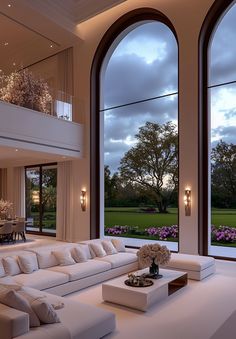 a living room filled with white furniture and large windows overlooking a lush green field at dusk