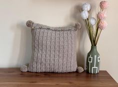 a knitted pillow sitting on top of a wooden table next to a vase with flowers