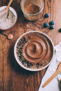 a wooden table topped with two bowls filled with chocolate frosting and blueberries next to spoons