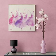 a vase with white flowers on a table next to a pink wall and a painting