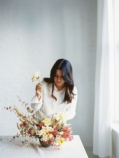 a woman sitting at a table with flowers in front of her and holding a vase