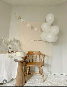 a table topped with a cake next to a wooden chair and white balloons hanging from the ceiling