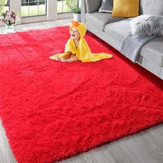 a baby is laying on the red rug in front of a gray couch and window