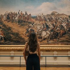 a woman standing in front of a painting with horses and men on it's walls