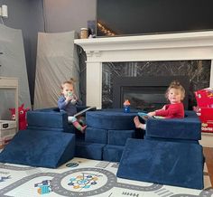 two children sitting on blue couches in front of a fire place with toys around them