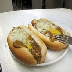 two hotdogs with chili and cheese on a white plate next to a fork