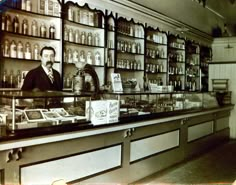 an old photo of a man behind the counter in a store with shelves full of bottles