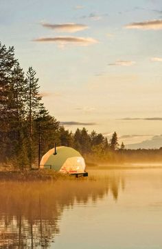 a tent is sitting on the shore of a lake with fog in the air and trees around it