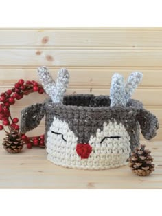 a crocheted basket with a reindeer face and pine cones on the side, sitting on a wooden surface