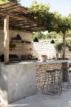 an outdoor kitchen with bar stools under a pergolated roof and stone walls