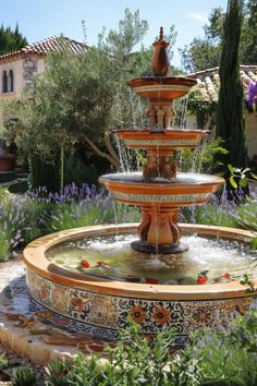 a fountain in the middle of a garden with purple flowers and greenery around it